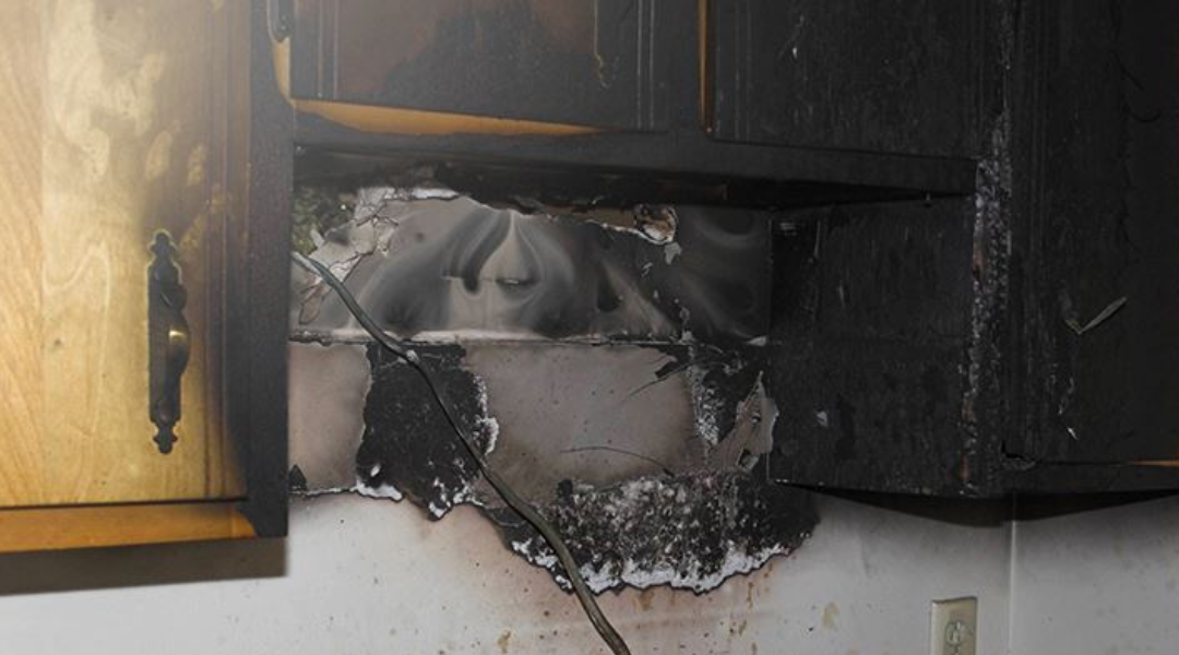 Close-up of kitchen cabinets showing severe fire damage, charred surfaces, and smoke stains near an electrical wire.