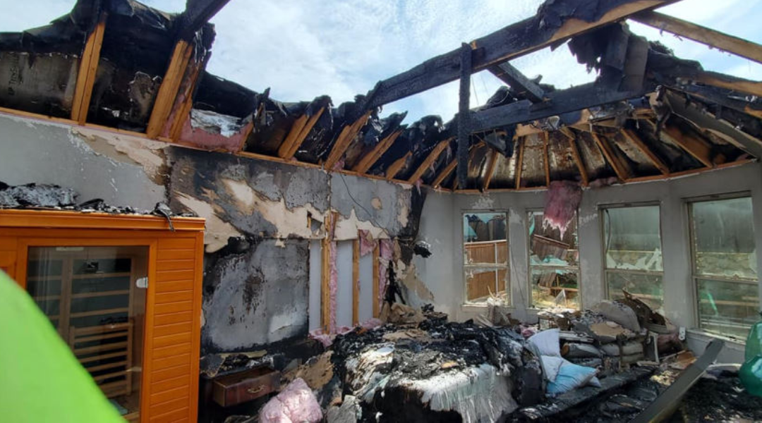 Interior of a fire-damaged room with a charred ceiling, burned walls, and debris strewn across the floor. Light shines through broken windows.