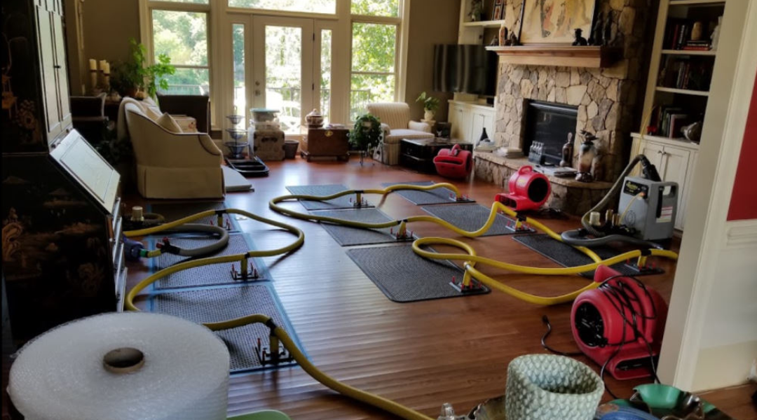A flooded living room with drying mats, hoses, and fans; furniture is shifted, and items are cluttered nearby. Sunlight filters through windows.