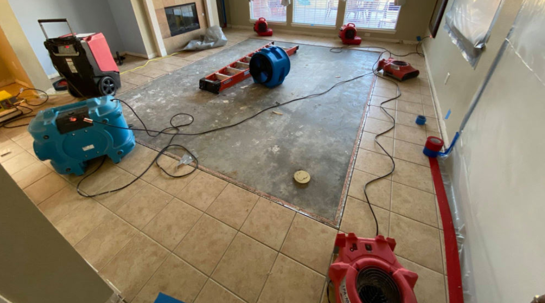 A partially renovated room with drying equipment, including fans and a dehumidifier, on tiled flooring and exposed subfloor.